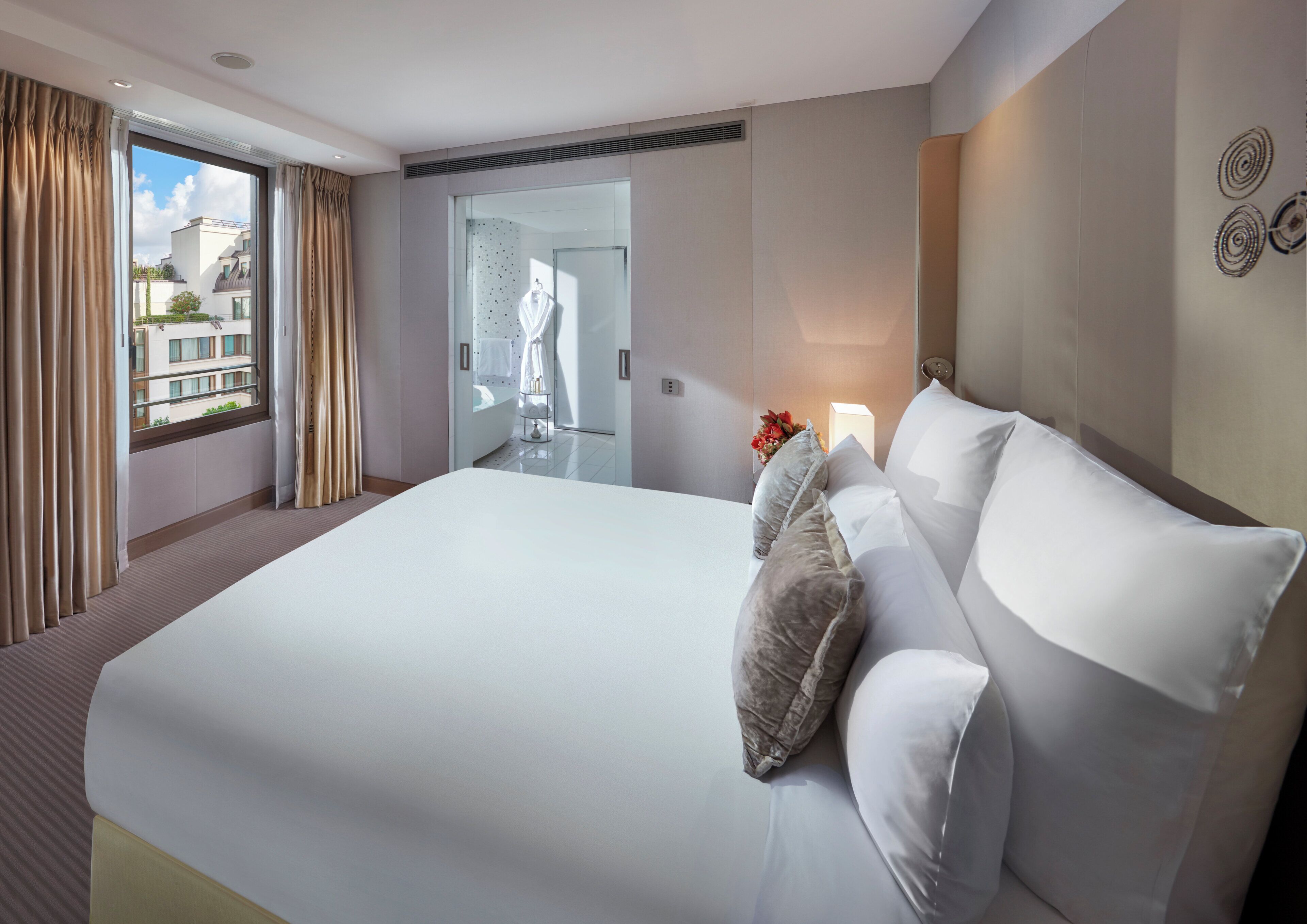A large bed with white linens and multiple pillows, a bedside table with a vase of flowers, a window with curtains, and a view of the outside buildings.