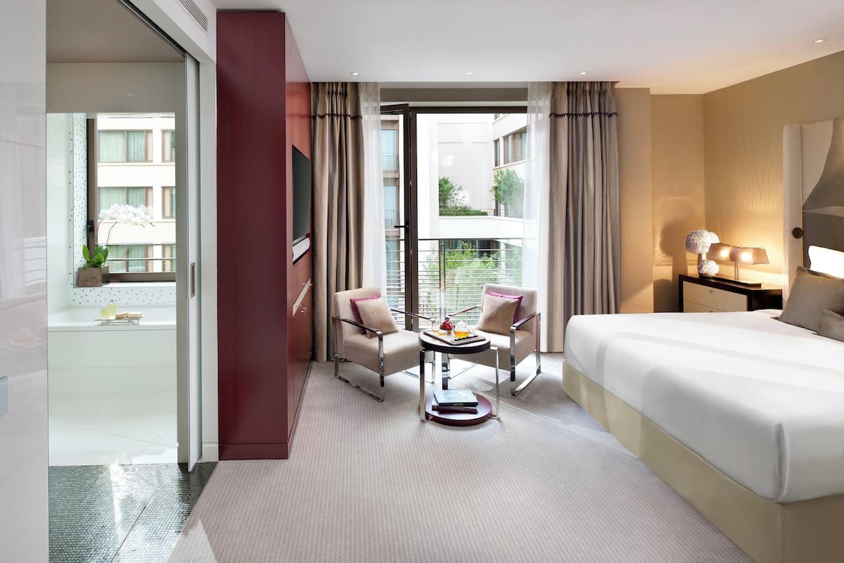 A hotel room with a bed, two armchairs, a small table, a TV, and a balcony with curtains.