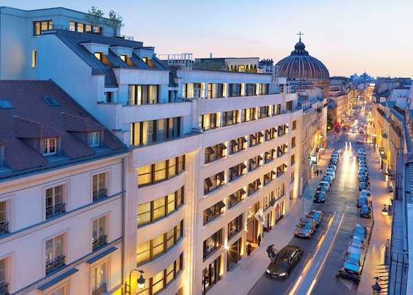 Un hotel de varios pisos con balcones, un edificio en forma de cúpula al fondo y una calle concurrida con coches estacionados.