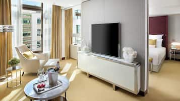 Una habitación de hotel moderna con un televisor de pantalla plana grande, una cama, un área de descanso con una silla y una mesa pequeña, y un balcón con plantas.