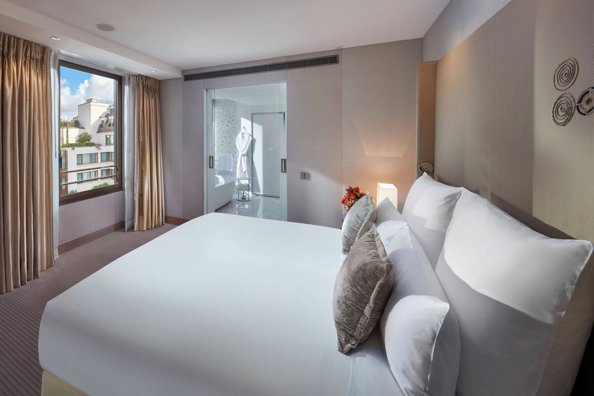 A large bed with white linens and multiple pillows, a bedside table with a vase of flowers, a window with curtains, and a view of the outside buildings.