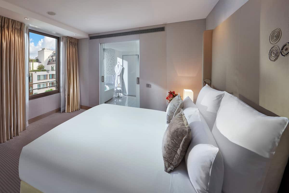 Ein großes Bett mit weißen Leinentüchern und mehreren Kissen, ein Nachttisch mit Blumen in einer Vase, ein Fenster mit Vorhängen und Blick auf die äußeren Gebäude.