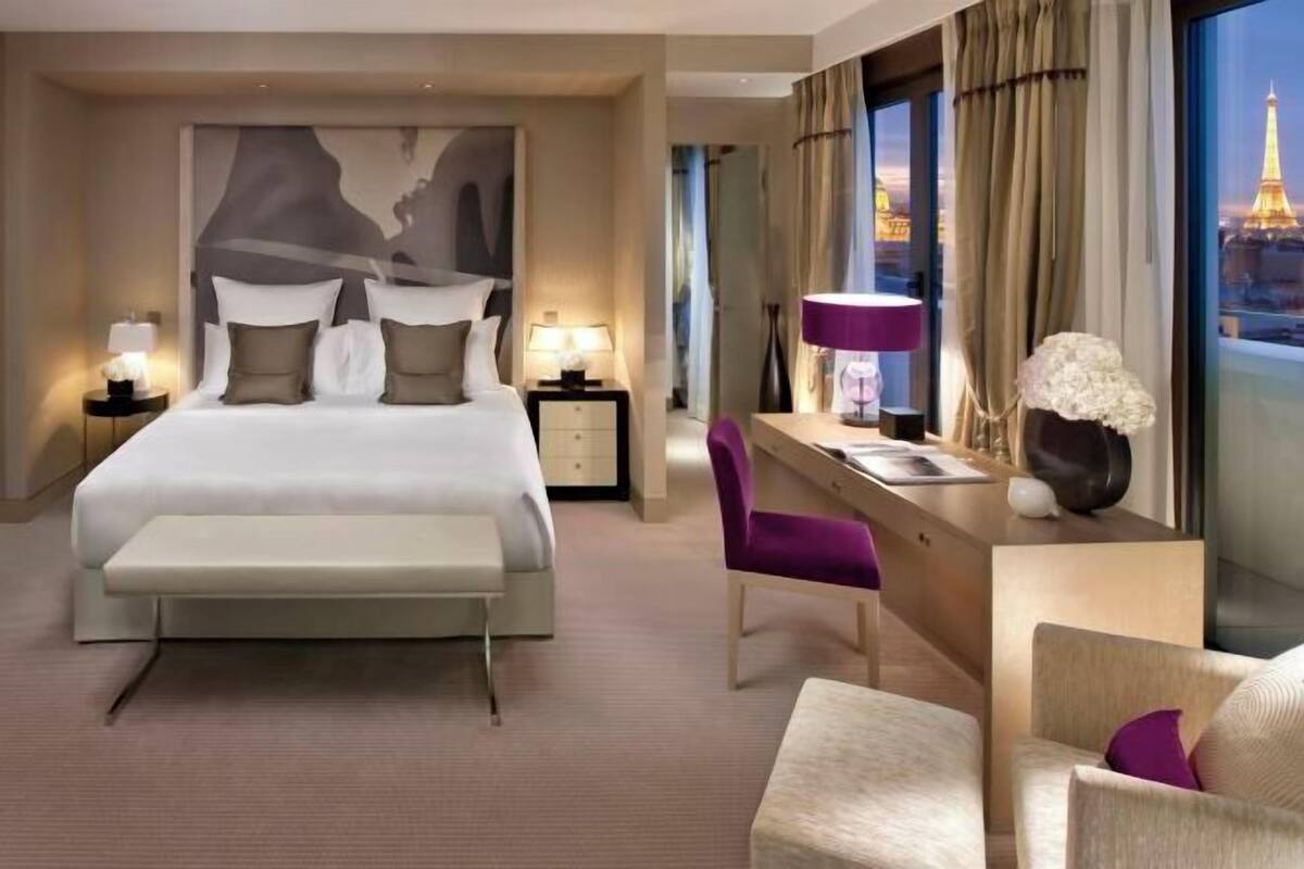 A hotel room with a large bed, bedside tables with lamps, a desk with a chair, and a view of the Eiffel Tower.