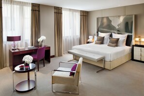 A hotel room with a large bed, a desk, a chair, a small table, and a nightstand.