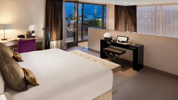 A modern hotel room with a large bed, a desk, and a view of the cityscape through floor-to-ceiling windows.