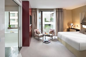 A hotel room with a bed, two armchairs, a small table, a TV, and a balcony with curtains. - Mandarin Oriental, Paris (Paris)