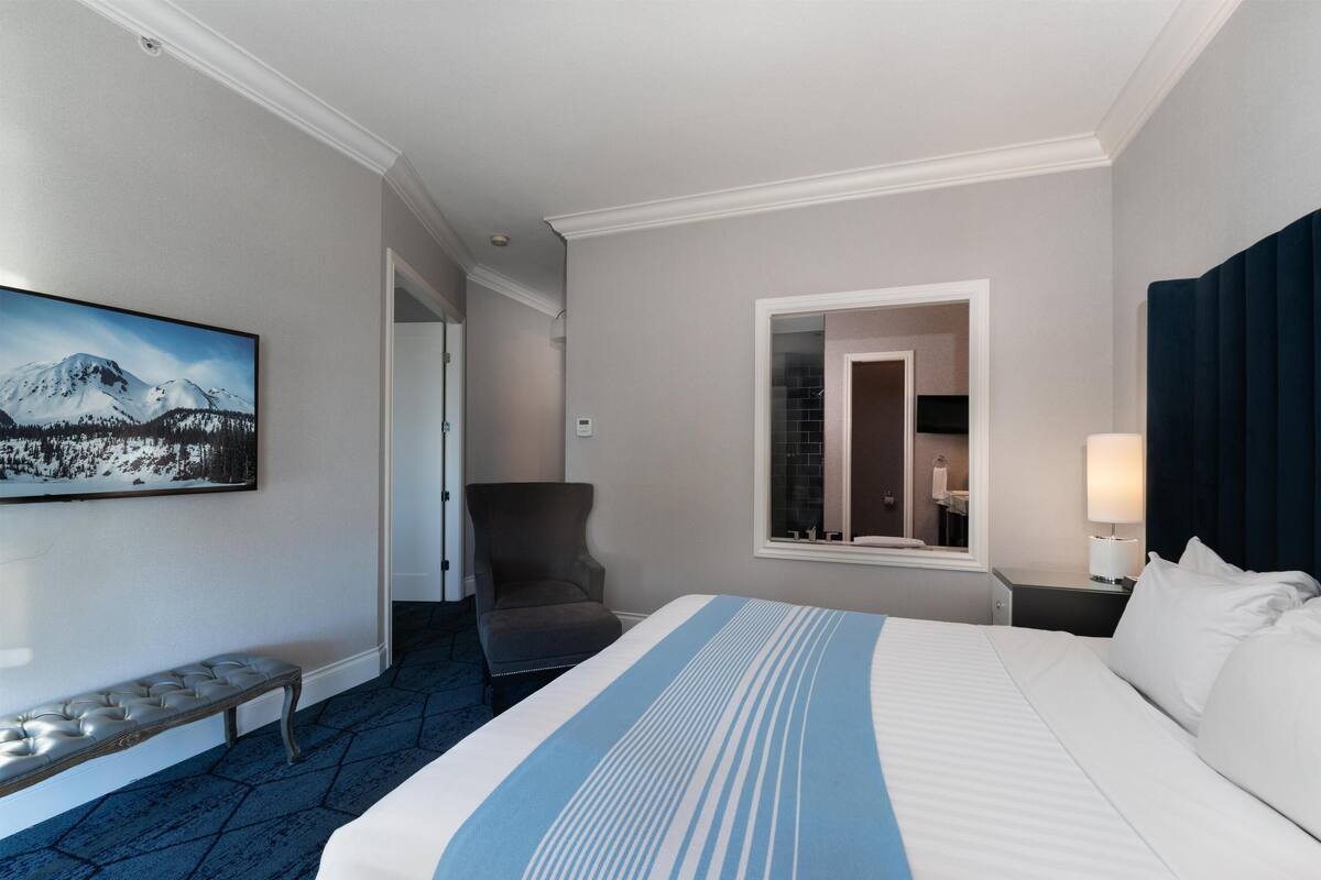 Quarto de hotel com cama, um banquinho, uma cadeira, janela com vista para montanha e um quadro na parede.