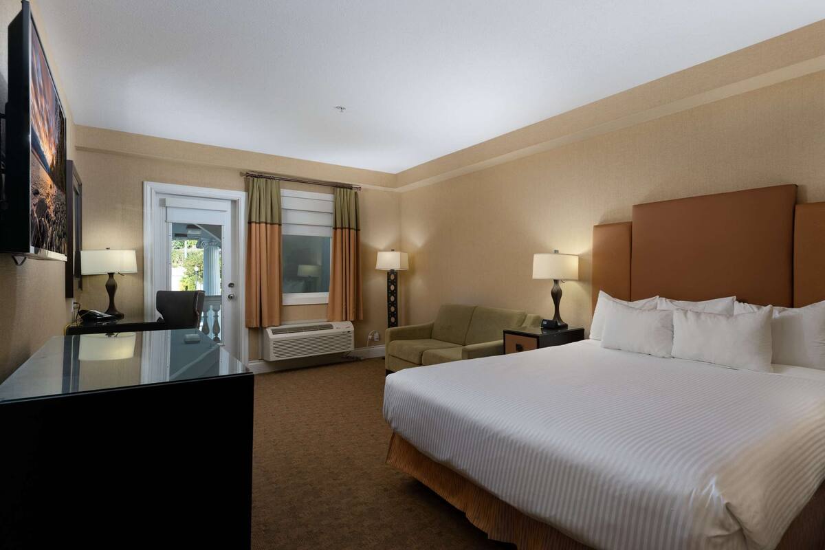 Quarto de hotel com uma cama grande, duas mesas de cabeceira com luminárias, uma escrivaninha com cadeira, uma televisão na parede e uma janela com cortinas.