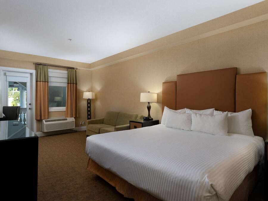 Habitación de hotel con una cama grande, dos mesitas de noche con lámparas, un escritorio con silla, un televisor montado en la pared y una ventana con cortinas.