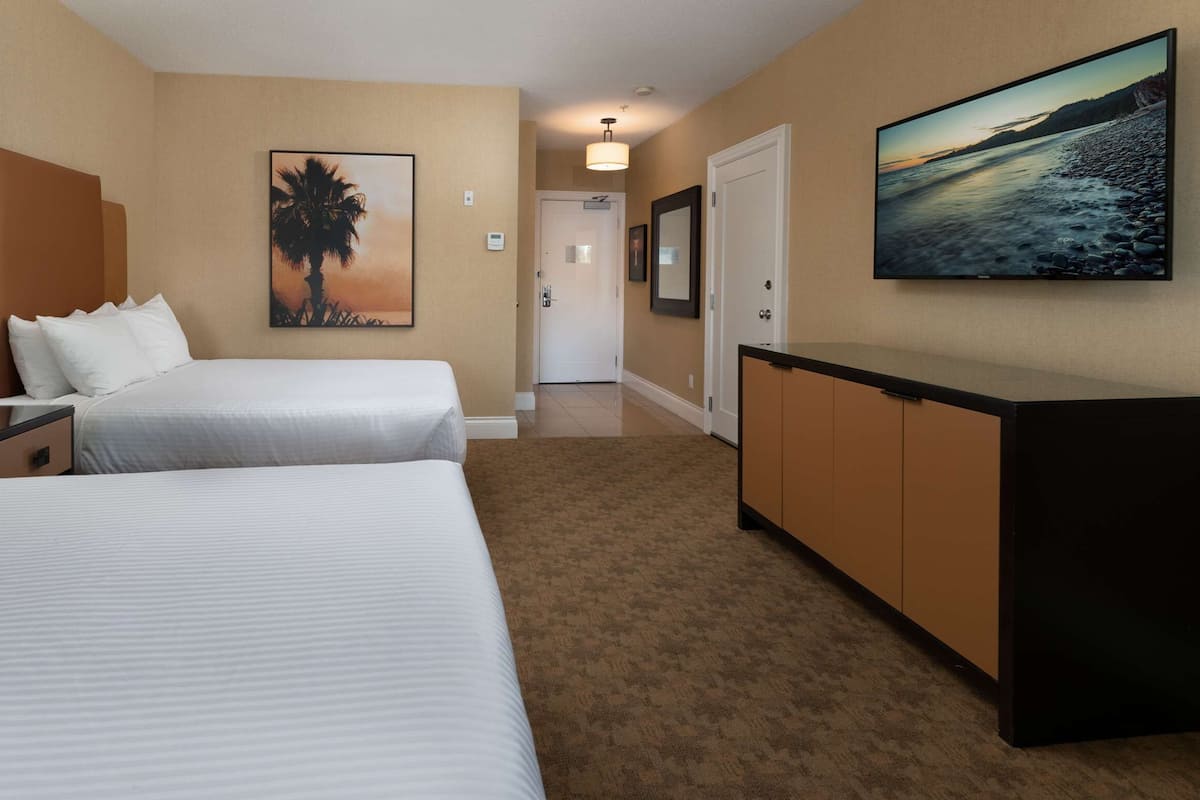Quarto de hotel com duas camas, uma escrivaninha, uma luminária, um quadro de palmeira e uma televisão na parede.