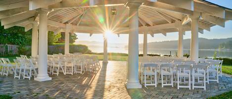 A covered outdoor seating area with white chairs arranged in rows, a pergola structure, and a view of a body of water in the background.