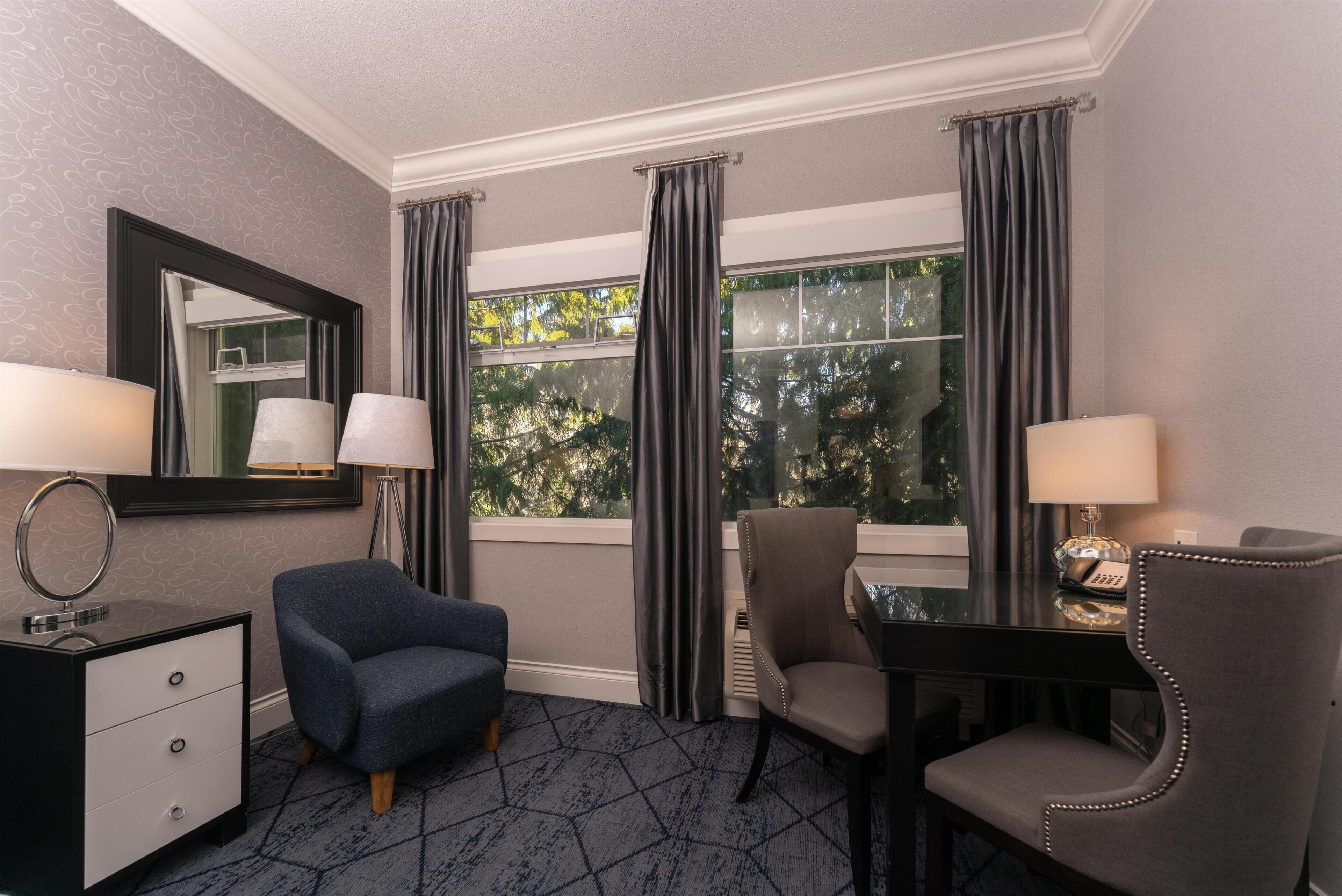 A hotel room with a large window, two chairs, a desk, a mirror, and a dresser.