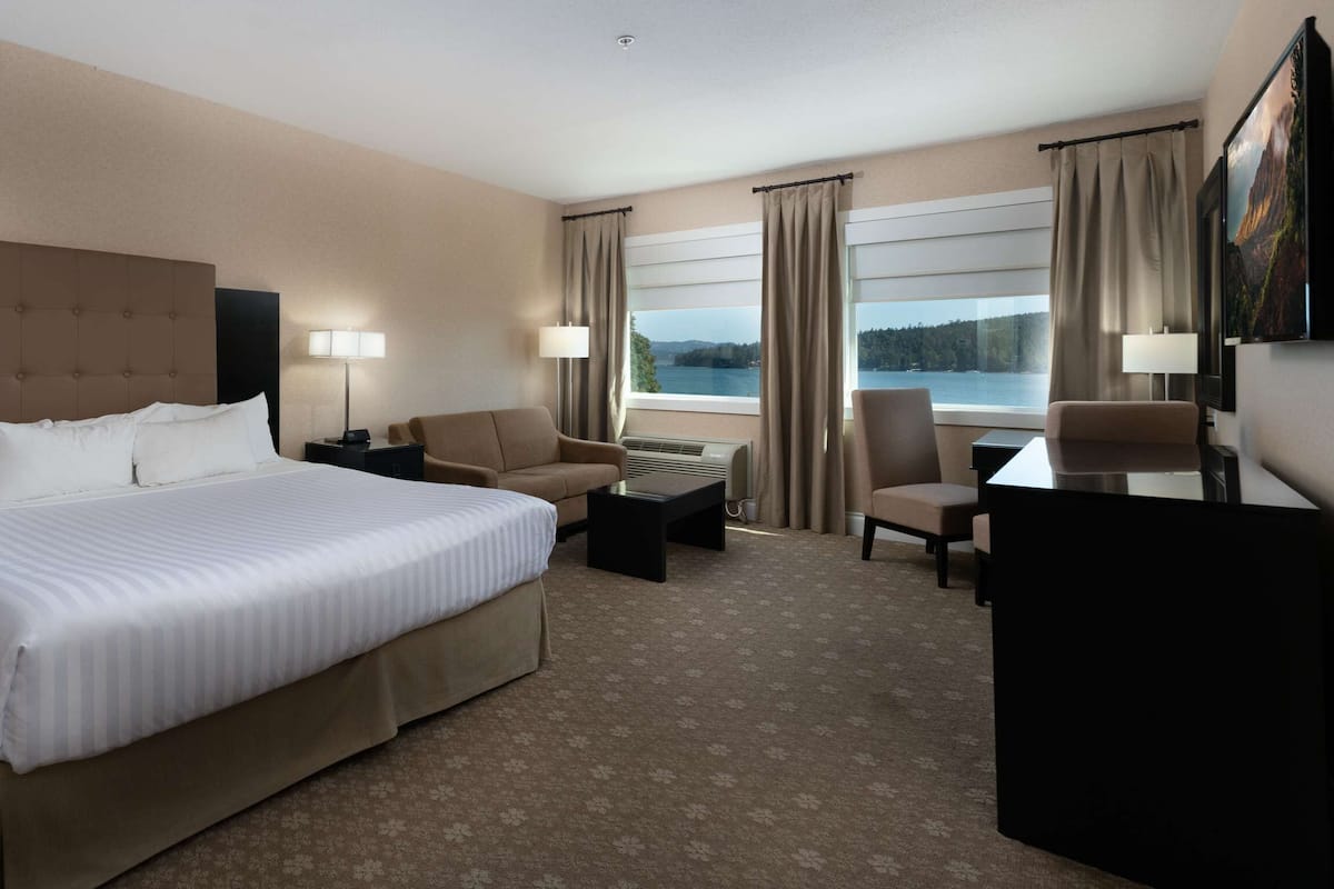 Quarto de hotel com uma cama grande, duas poltronas, uma escrivaninha e vista para um lago.
