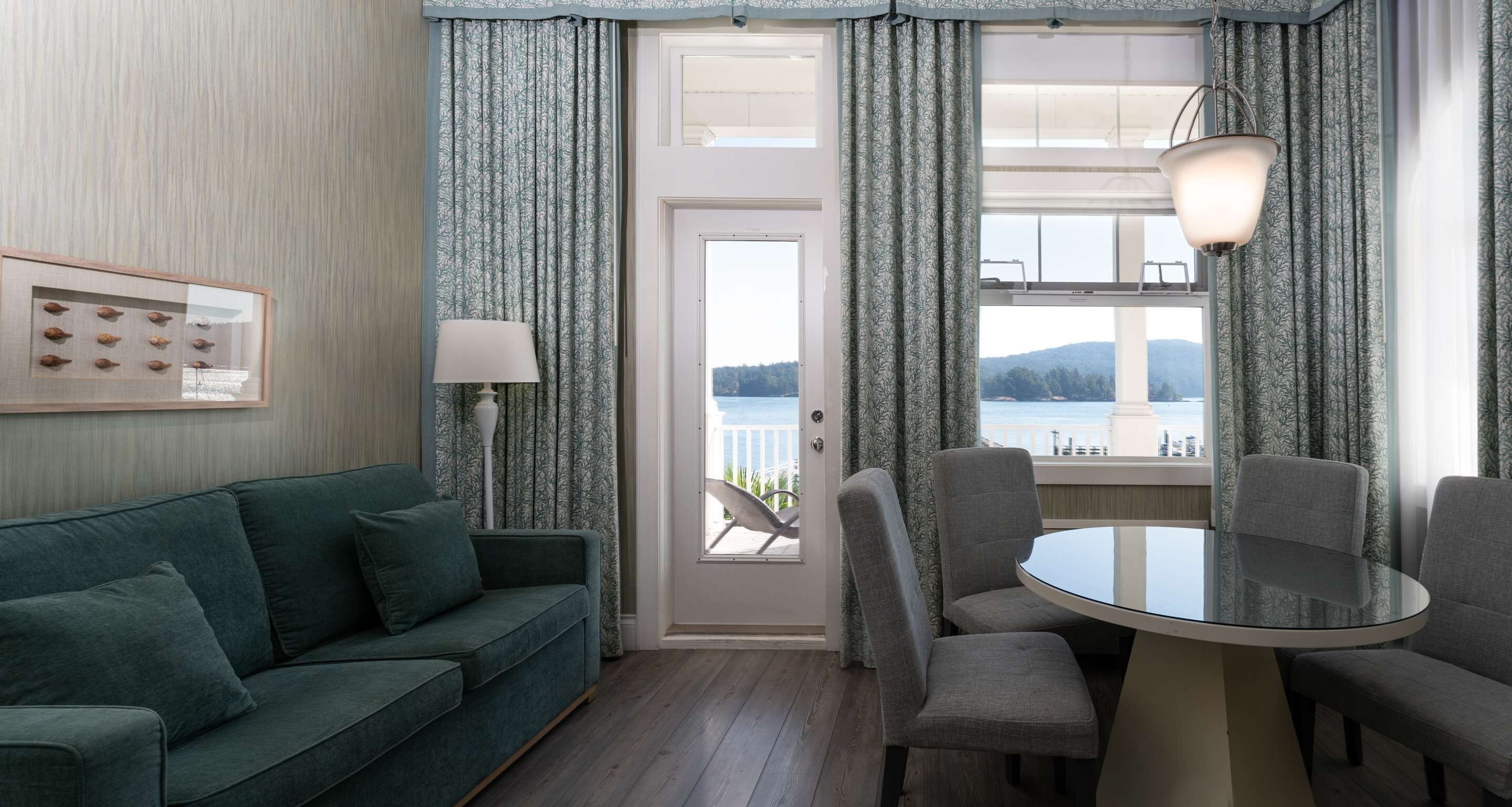 Una sala de estar moderna con un sofá verde, una mesa redonda de vidrio y sillas grises. Una puerta da a un balcón con vistas al agua y las colinas.