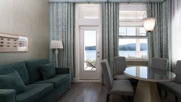 Una sala de estar moderna con un sofá verde, una mesa redonda de vidrio y sillas grises. Una puerta da a un balcón con vistas al agua y las colinas.