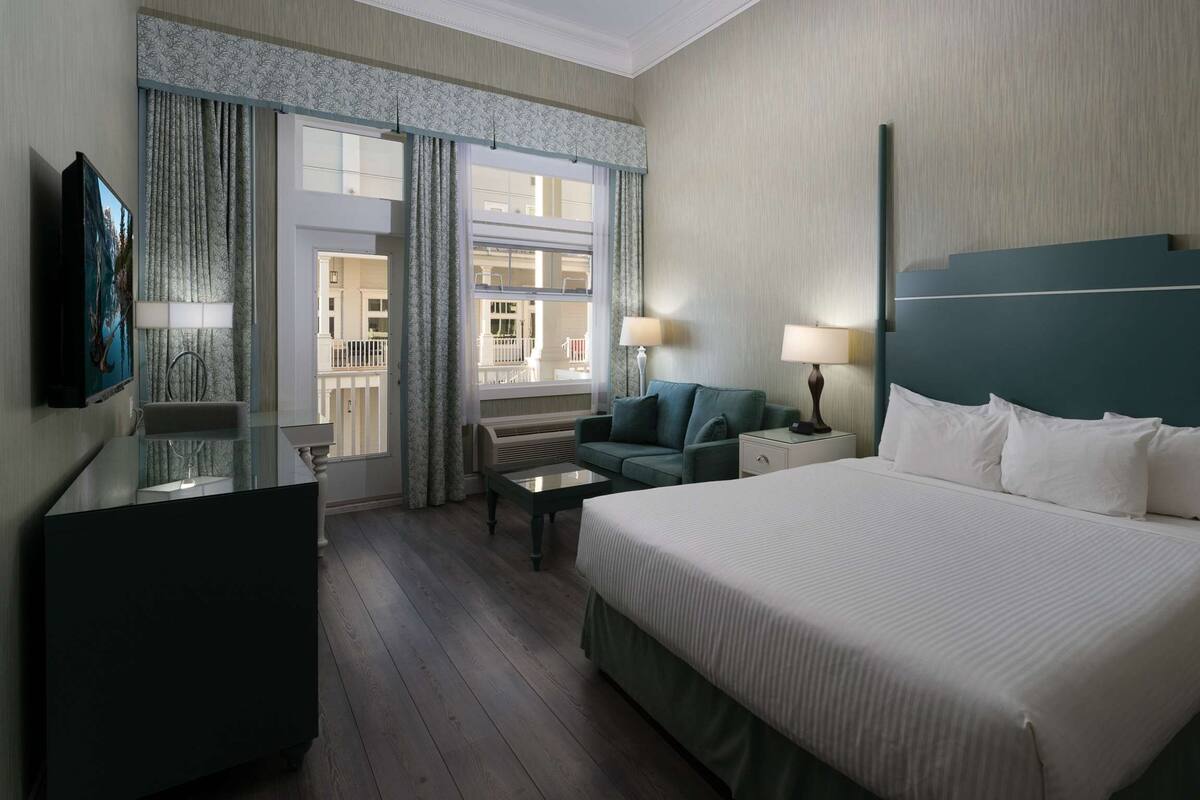Quarto de hotel com uma cama grande, uma escrivaninha, uma cadeira, uma TV e uma janela com cortinas.