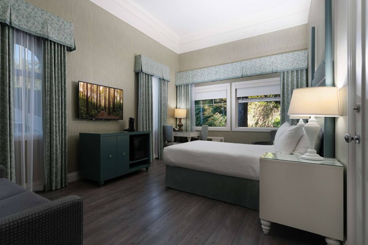 Quarto de hotel com cama, escrivaninha, cadeira, televisão e janela com cortinas.