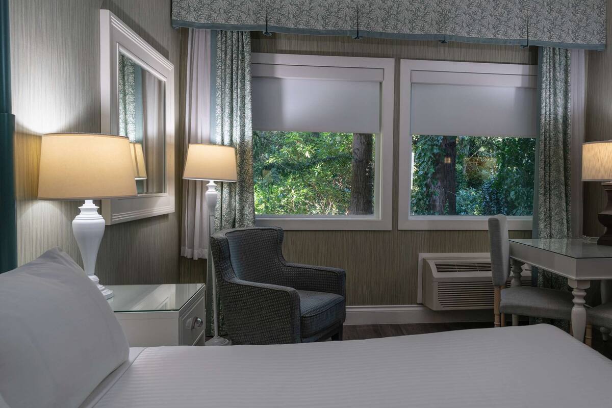 Quarto de hotel com uma cama, duas cadeiras, uma mesa e uma janela com vista para vegetação.