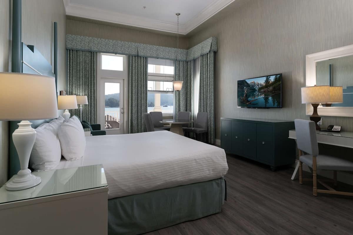 Quarto de hotel com cama, escrivaninha, cadeira, cômoda e uma janela grande com cortinas.