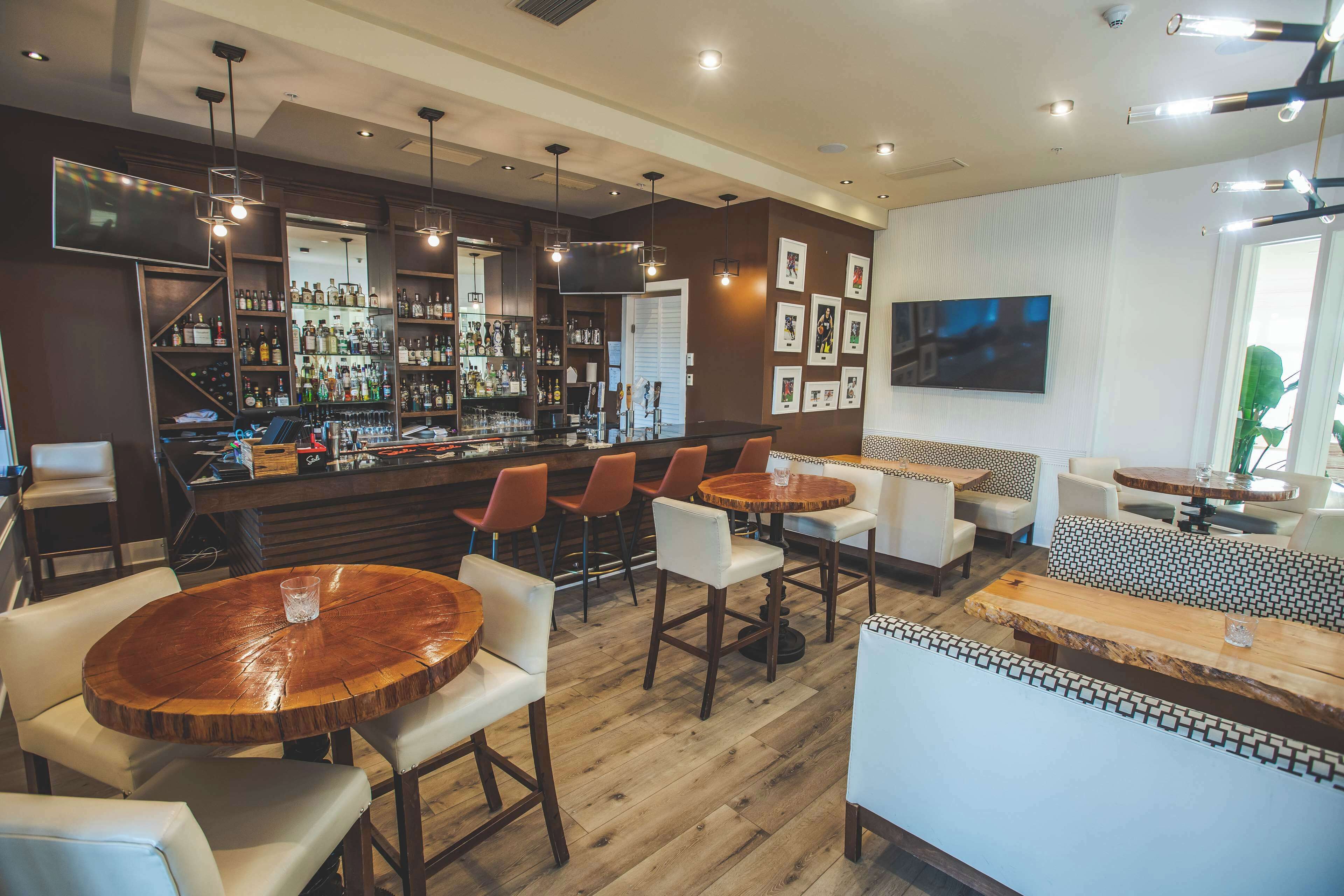 Una zona de bar con mesas y sillas de madera, un televisor montado en la pared y una variedad de botellas en estantes.