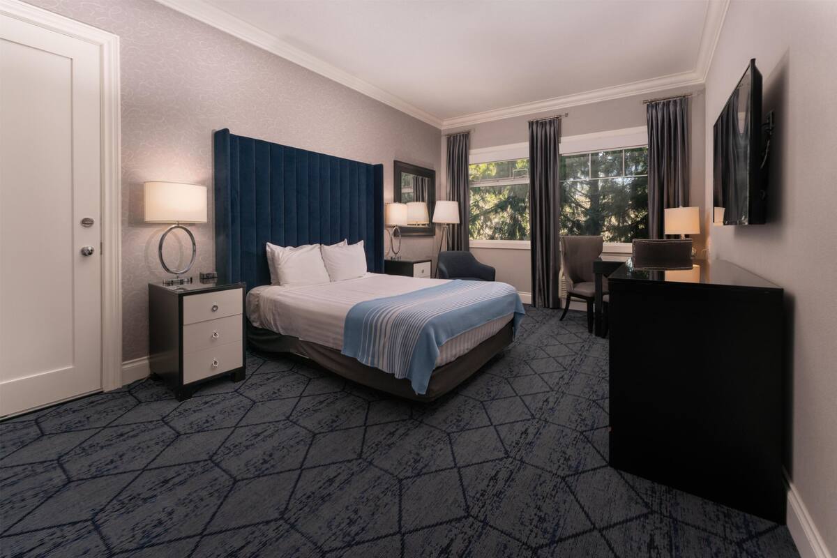 Quarto de hotel com cama, mesas de cabeceira, escrivaninha, cadeira, televisão e janela com cortinas.