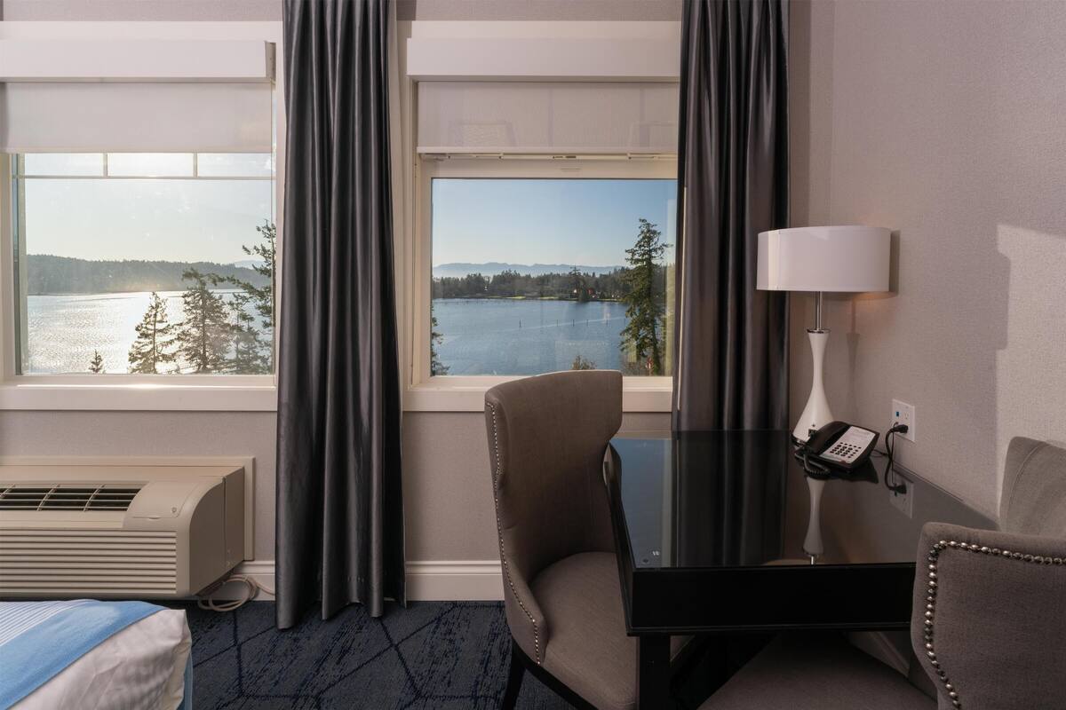 Quarto de hotel com cama, escrivaninha com telefone, luminária e vista para um lago e montanhas.
