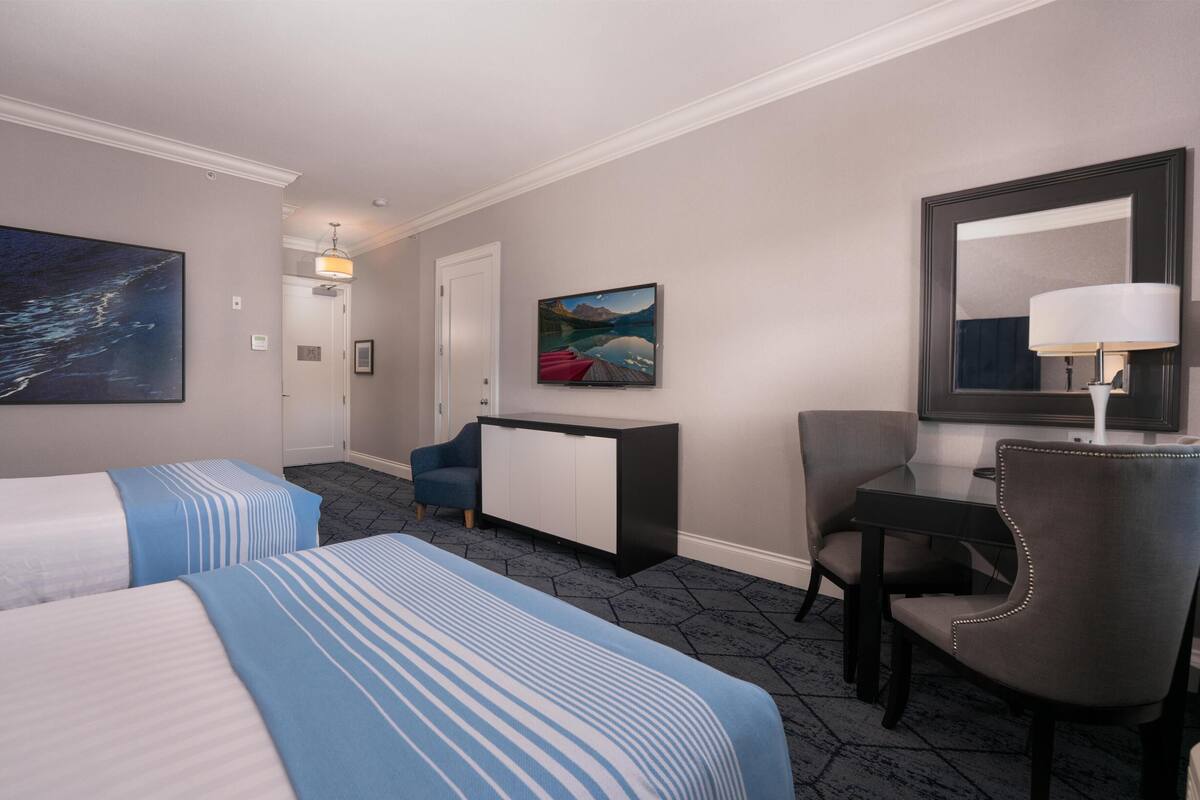 Quarto de hotel com duas camas, uma escrivaninha, uma cadeira, um espelho grande e obras de arte nas paredes.