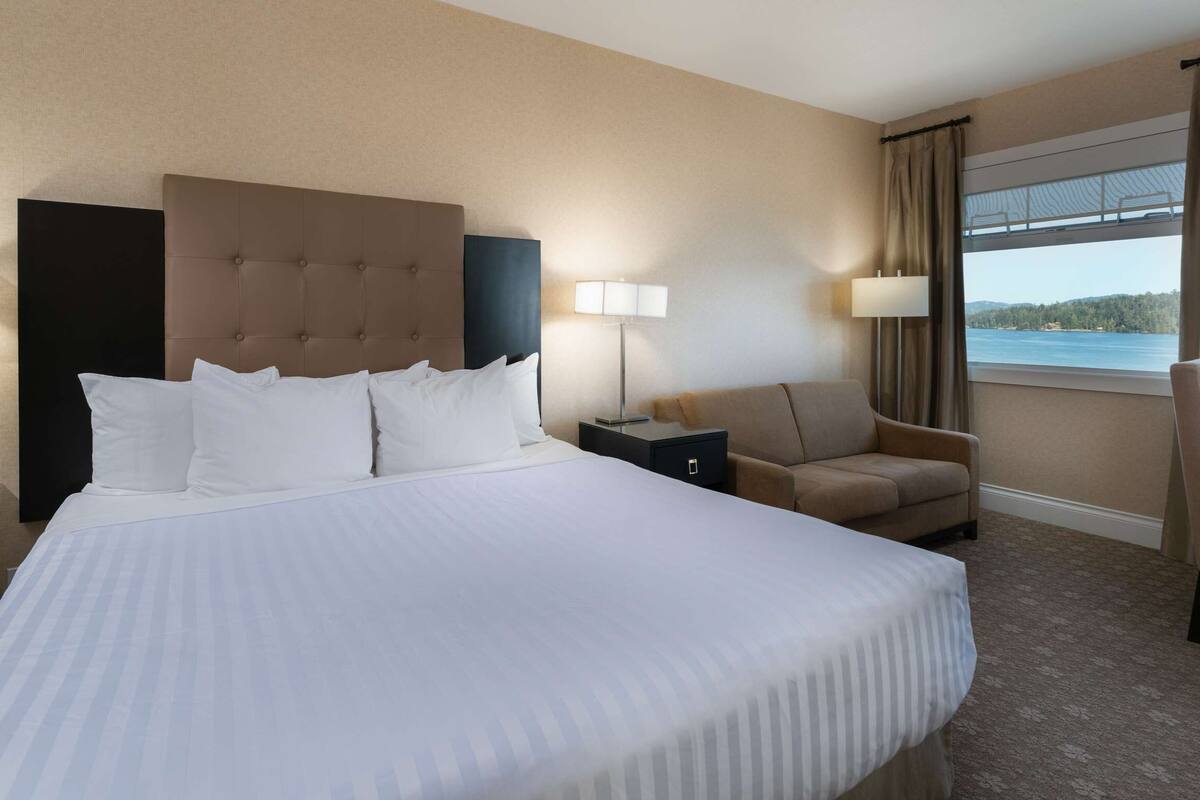 Quarto de hotel com uma cama grande, duas poltronas, uma escrivaninha e uma janela com vista para a água.