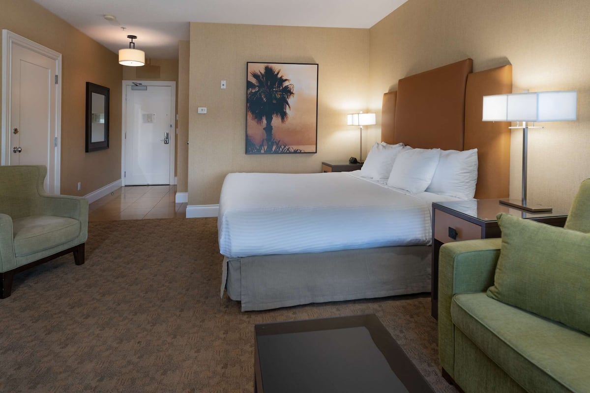 Quarto de hotel com cama, duas mesinhas de cabeceira, um criado-mudo, uma poltrona verde, um sofá verde, um quadro na parede e uma porta.