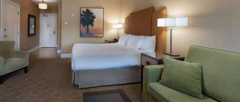A hotel room with a bed, two bedside tables, a nightstand, a green armchair, a green sofa, a framed picture on the wall, and a door.