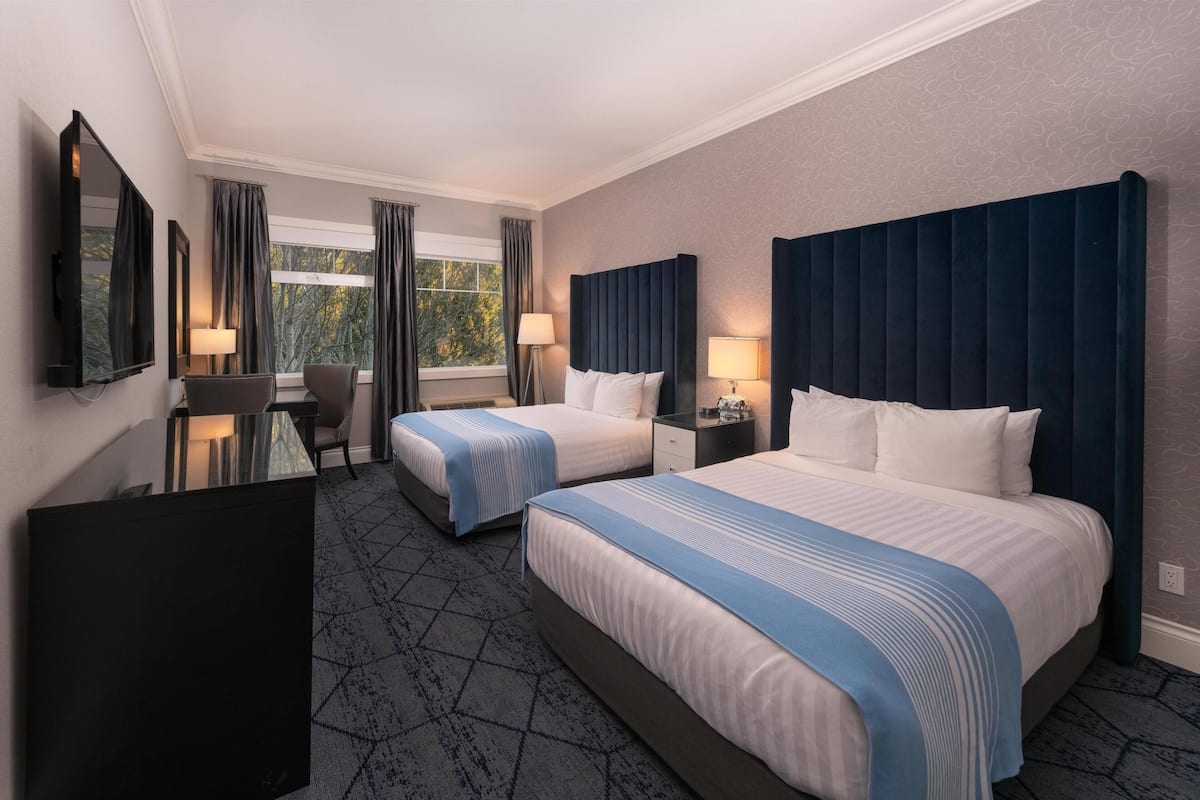 Quarto de hotel com duas camas, uma mesa, uma cadeira, uma televisão e uma janela com cortinas.
