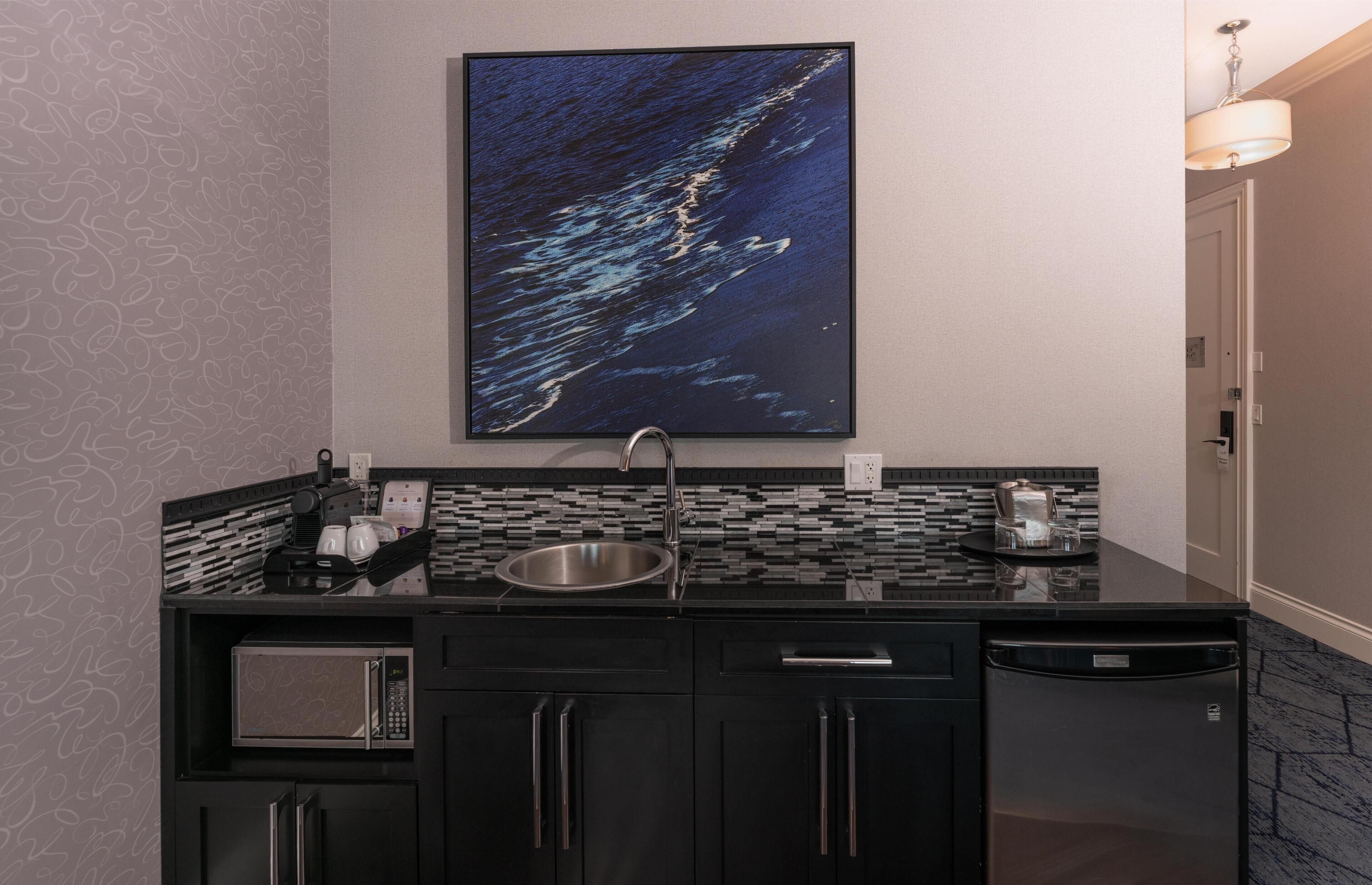 A modern kitchen with a microwave, sink, and dishwasher, a large artwork above the counter, and a patterned wall.