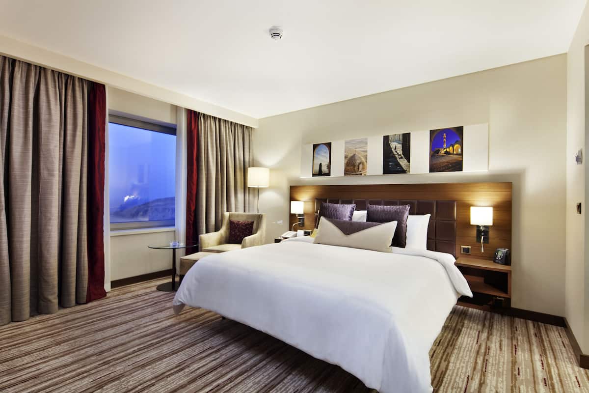A hotel room with a large bed, a desk, a chair, a nightstand, and a window with curtains.