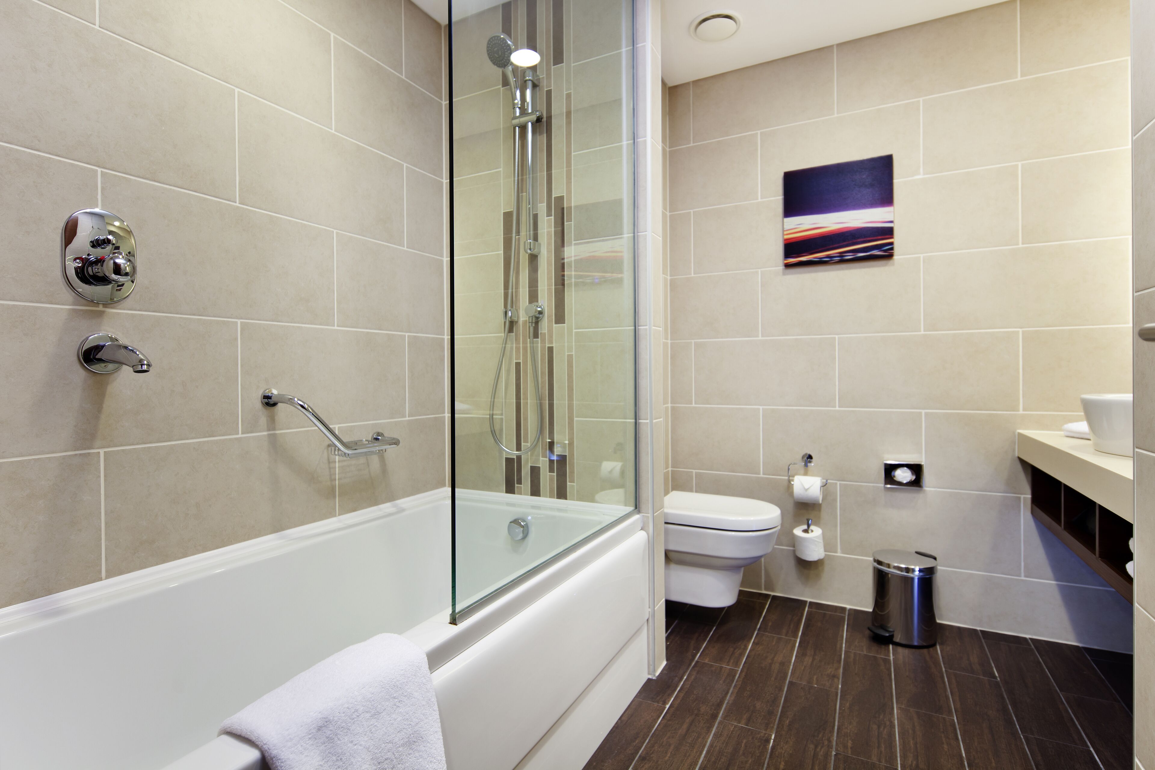A modern bathroom with a glass-enclosed shower, a white bathtub, a toilet, and a trash can.