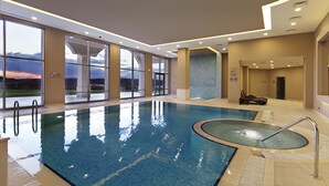 An indoor swimming pool with clear glass walls, a view of the sunset, and a lounge area with chairs. - Hilton Garden Inn Mardin (Mardin)