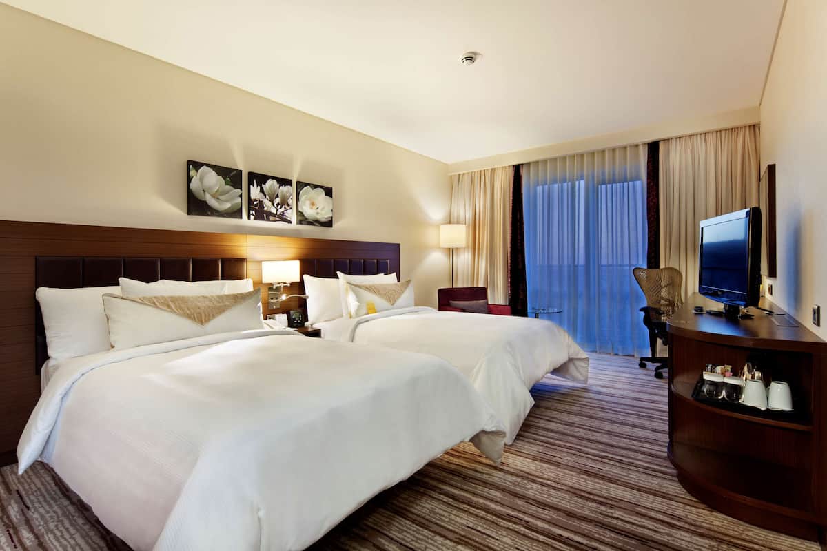 A hotel room with two beds, a TV, a desk, and a large window with curtains.