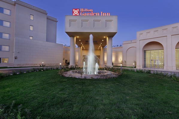 Um Hilton Garden Inn com uma fonte central, colunas imponentes e uma entrada bem iluminada.