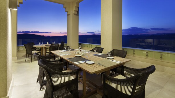 A dining area with a table set for four, surrounded by wicker chairs, overlooking a scenic view of hills during twilight. - Hilton Garden Inn Mardin (Mardin)