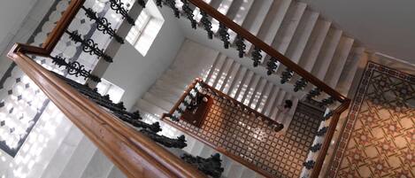 A grand staircase with a wooden handrail and ornate metal balustrade, leading to a landing with a patterned floor.
