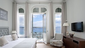 A hotel room with a large bed, a chair, a TV, and a view of the sea through open windows. - Poseidonion Grand Hotel (Spetses)