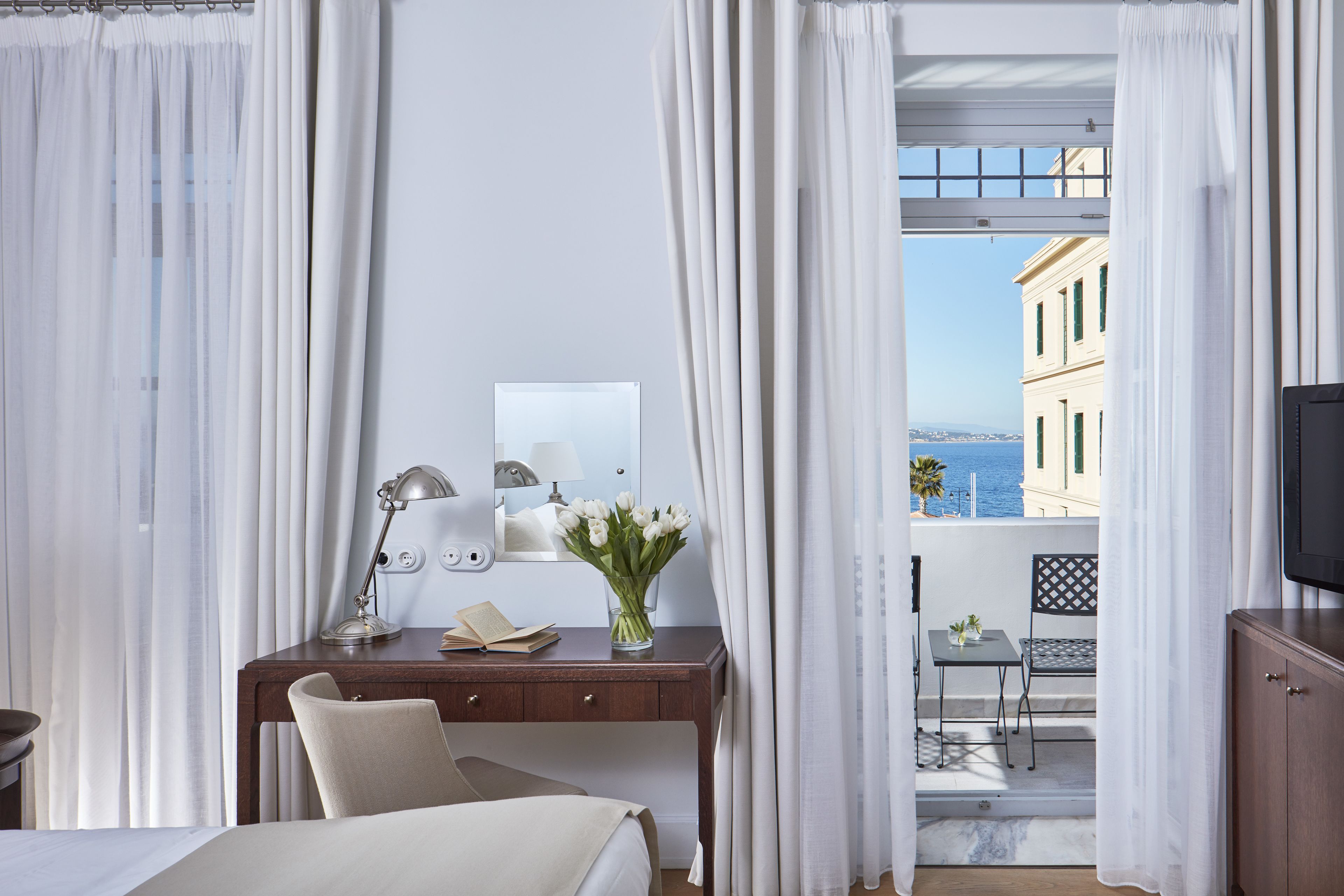 Ein Hotelzimmer mit einem Bett, einem Schreibtisch mit Lampe, einem Spiegel, einer Vase mit Blumen und Blick auf das Meer und die Gebäude.