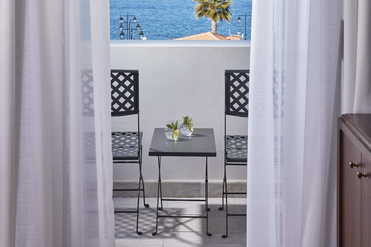 Ein Balkon mit Tisch und Stühlen, der Blick auf die Küste mit einer Palme und dem Meer freigibt.