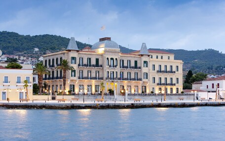 A large, multi-story building with a prominent dome and a flag on top, situated by a waterfront with a stone wall and benches along the promenade.