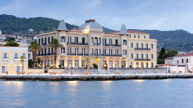 A large, multi-story building with a prominent dome and a flag on top, situated by a waterfront with a stone wall and benches along the promenade. - Poseidonion Grand Hotel (Spetses)