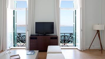 Quarto com televisão sobre um armário de madeira, um banco, uma mesa com um livro e um abajur.