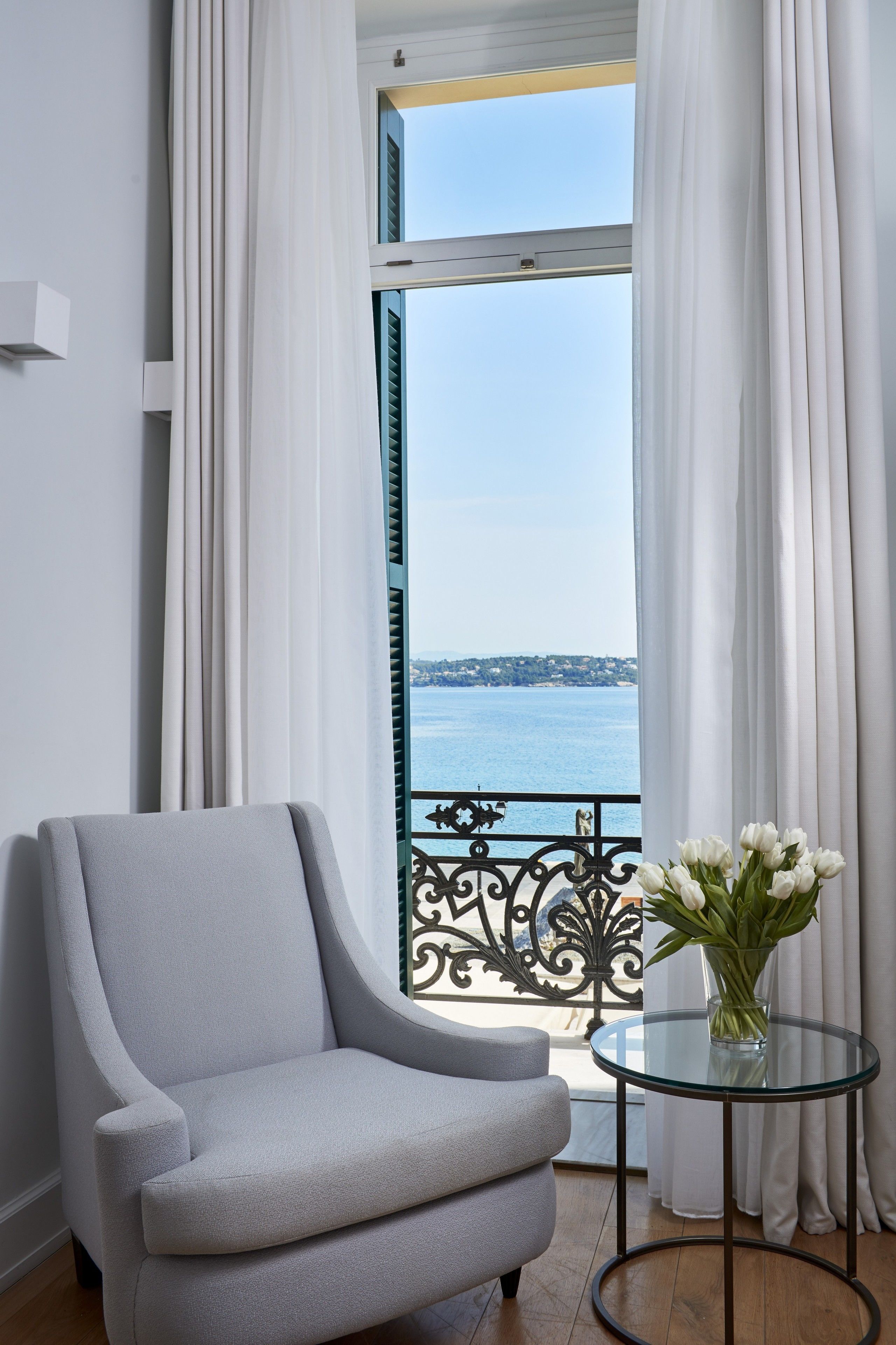 Una habitación con vista al mar, un sillón gris, una mesa de vidrio con un jarrón de flores y cortinas blancas.