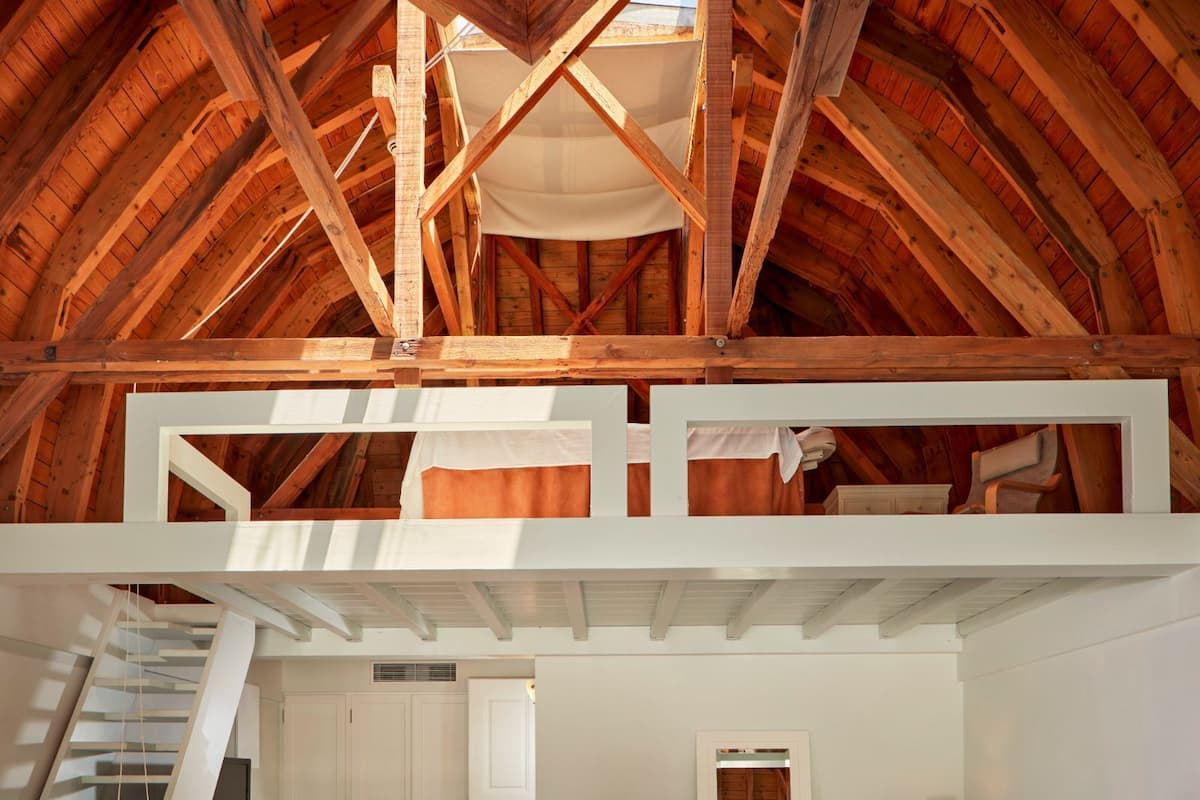 Ein geräumiges Schlafzimmer im Loft-Stil mit hohen Decken, Holzbalken, einer eingebauten Wendeltreppe, einem großen Bett, einem Sessel und einem Schreibtisch.