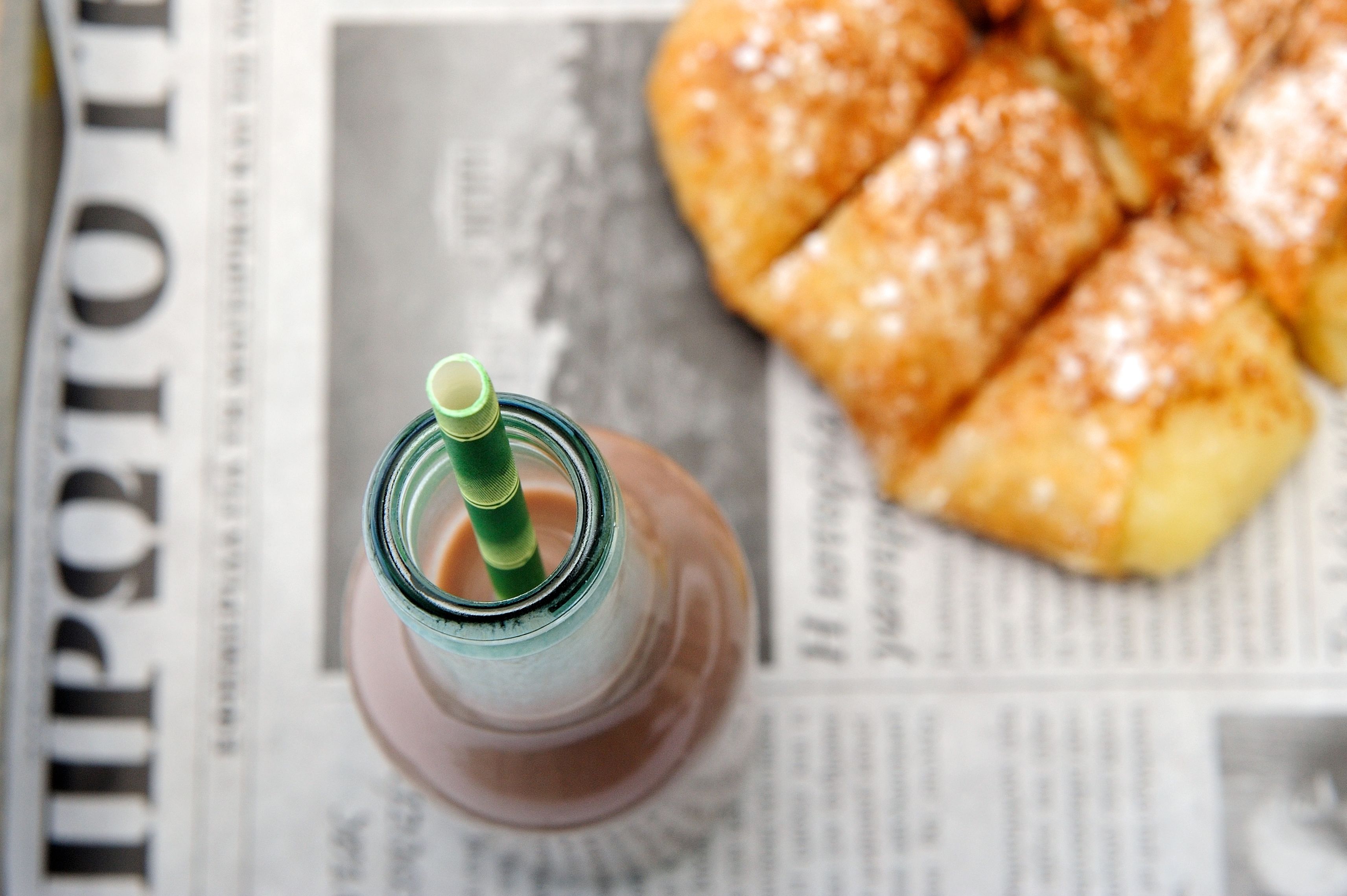 Une bouteille en verre avec une paille verte, posée sur un journal à côté d’un croissant.