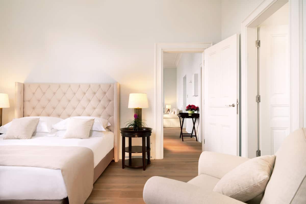 Ein Hotelzimmer mit einem großen Bett, einem Nachttisch mit Lampe, einem Sessel und Blick auf den Flur.