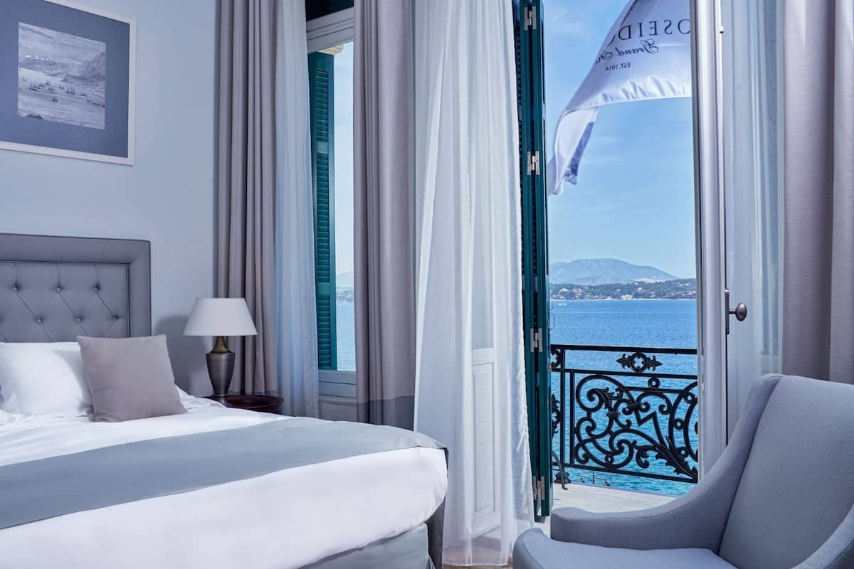 Ein Hotelzimmer mit Bett, Nachttisch, Lampe und Meerblick von einem Balkon mit Geländer.